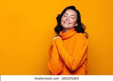 Delighted positive brunette woman smiling, wear high neck knitted orange sweater, has good mood, dreamily closed her eyes, isolated on studio orange background with mockup space. Autumn season.