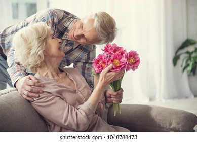 Delighted retired female sitting on sofa with flowers in hands. Old man standing behind and looking at her with smile - Powered by Shutterstock