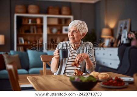 A delighted senior adult woman sitting at a dining table and looking through window while enjoying a delicious croissant and a cup of coffee.
