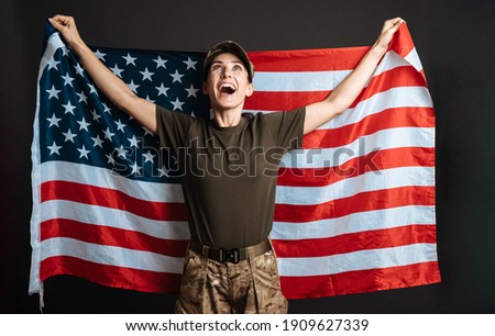 Delighted soldier woman smiling while posing with american flag isolated over black background