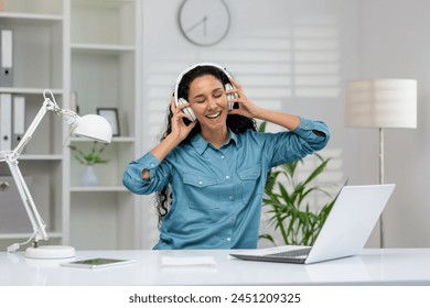 A delighted woman in a blue shirt listens to music with headphones, smiling broadly as she enjoys a break at her clean, contemporary office workspace. - Powered by Shutterstock