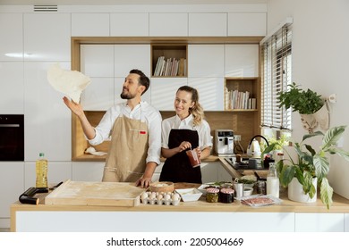 Delighted Woman Chef With Man Chef Cooking Baking Preparing Dough For Domestic Pizza In Modern Light Kitchen. Pizza Ingredients On Wooden Surface Eggs Flour Olives Asparagus Mushrooms Tomato Sauce.