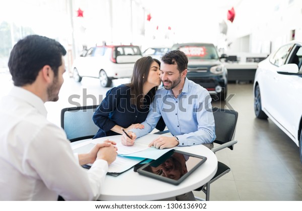 Delighted woman kissing man signing purchase\
and sale agreement in car\
dealership
