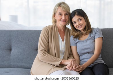 Delighted Young Asian Girl Hugging Happy Middle Aged Foster Mother Sitting On Sofa While Spending Time Together At Home, Adopted Child And Mother In Law Conceppt.