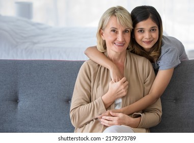 Delighted Young Asian Girl Hugging Happy Middle Aged Foster Mother Sitting On Sofa While Spending Time Together At Home, Adopted Child And Mother In Law Conceppt.