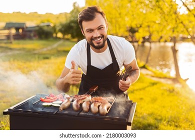 Delighted young ethnic bearded man in black apron smiling at camera and showing thumb up gesture, while preparing delicious grilled sausages and vegetables during picnic in park at sunset - Powered by Shutterstock