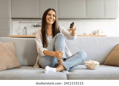 Delighted young female with bowl of popcorn at home comfortably watching her favorite tv shows, smiling woman holding remote in hand, relaxing on plush couch in living room, copy space