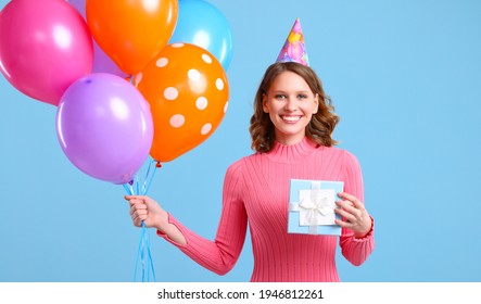 Delighted Young Female Smiling And Looking At Camera While Showing Bunch Of Colorful Balloons And Wrapped Birthday Present Against Blue Background