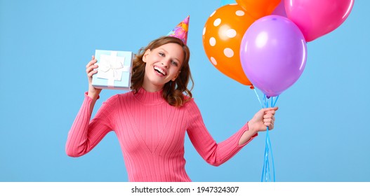 Delighted Young Female Smiling And Looking At Camera While Showing Bunch Of Colorful Balloons And Wrapped Birthday Present Against Blue Background