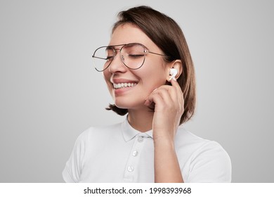 Delighted young female in stylish eyeglasses touching true wireless earbuds while listening to songs in studio against gray background - Powered by Shutterstock