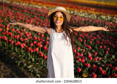 Delighted young female tourist in white dress straw hat and sunglasses, smiling and looking at camera while standing in tulip field during trip in Netherlands
