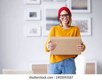 Delighted young female in trendy outfit smiling for camera and carrying cardboard box at home