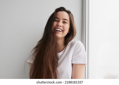 Delighted young lady with long dark hair in t shirt smiling happily and looking at camera, while sitting near window and leaning on white wall - Shutterstock ID 2188181337