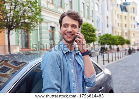 Delighted young male glancing aside while having phone conversation near car