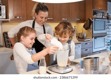 Delighted young mother in casual clothes and apron standing behind cute son and daughter weighting flour while helping to bake in kitchen