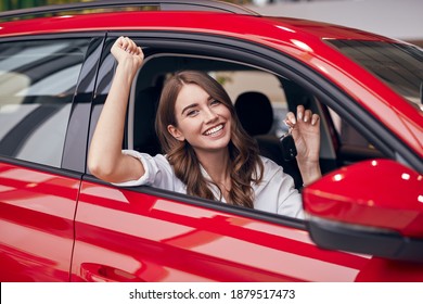 Delighted young woman showing key and celebrating success while sitting inside new red vehicle in dealership