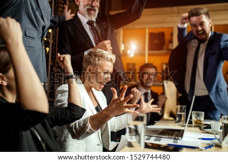 Delightful business people enthusiastically exulting in bright office, young woman happily raising hands smiling broadly at meeting