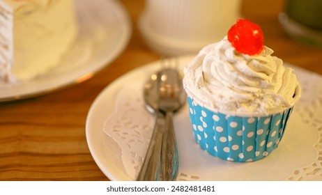 A delightful cupcake in a polka dot wrapper, topped with whipped cream and a cherry, sits on a doily-lined plate with a spoon, ready to enjoy. The scene exudes sweetness and charm. - Powered by Shutterstock