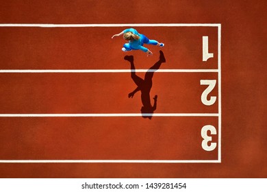 Delightful Girl In A Blue Sportswear And Sneakers Starts Her Sprint Run With A Long Jump On The Running Track Number 1 At The Stadium Outdoors. Sun Shines Onto Her Body. Top View Aerial Photo.