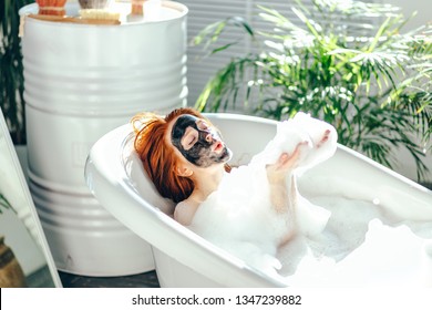 Delightful joyful caucasian young red-haired woman enjoying home spa in bathroom, lying in a foam bathtub with black clay face mask on face, playing with foam clouds - Powered by Shutterstock