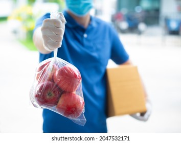 Deliver Man Wearing Medical Gloves And Face Mask In Blue Uniform Handling Apple Bag And Parcel Box Give To Female Costumer Postman And Express Grocery Delivery Service During Covid19.