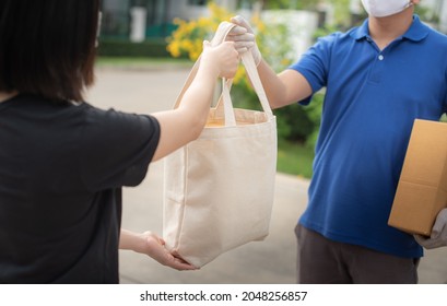 Deliver Man Wearing Medical Gloves And Face Mask In Blue Uniform Handling Bag Of Food And Parcel Box Give To Female Costumer Postman And Express Grocery Delivery Service During Covid19.