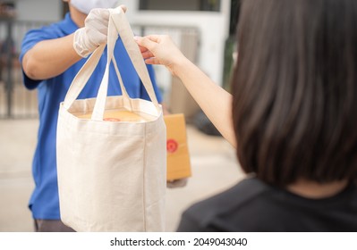 Deliver Man Wearing Medical Gloves And Face Mask In Blue Uniform Handling Bag Of Food And Parcel Box Give To Female Costumer Postman And Express Grocery Delivery Service During Covid19.