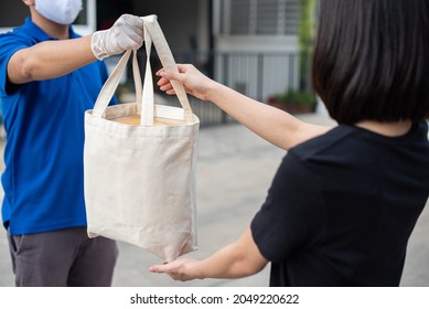 Deliver Man Wearing Medical Gloves And Face Mask In Blue Uniform Handling Bag Of Food And Parcel Box Give To Female Costumer Postman And Express Grocery Delivery Service During Covid19.