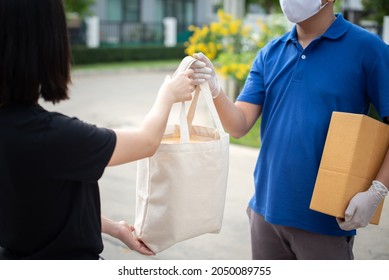 Deliver Man Wearing Medical Gloves And Face Mask In Blue Uniform Handling Bag Of Food And Parcel Box Give To Female Costumer Postman And Express Grocery Delivery Service During Covid19.