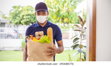 Delivery African man employee wearing face mask blue cap uniform mask hold paper bag packet organic food at home front door. Service quarantine pandemic. Express grocery delivery service courier. - Powered by Shutterstock