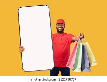 Delivery App. African American Courier Man Holding Big Cellphone With Empty Screen And Stretching Shopping Bags To Camera Over Yellow Background, Mockup