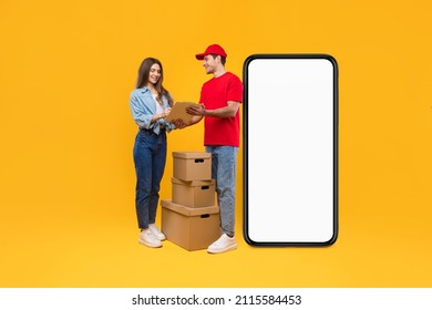 Delivery Application. Courier And Woman Near Huge Cellphone Blank Screen Signing Papers, Customer Receiving Parcel Boxes From Deliveryman Standing Over Yellow Studio Background. Mockup, Full Length