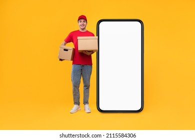 Delivery Application. Happy Courier Guy Near Huge Phone With Empty Screen Holding Cardboard Boxes Smiling To Camera Standing Over Yellow Studio Background. Collage, Mockup