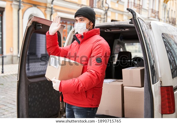 Delivery of big\
parcels by car with courier at home concept. Curier in medical mask\
waving and holding cardboard box near front door while standing at\
the street. Stock\
photo