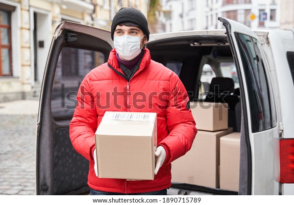 Delivery of big parcels by car\
with courier at home concept. Curier in medical mask holding\
cardboard box near front door while standing at the street. Stock\
photo