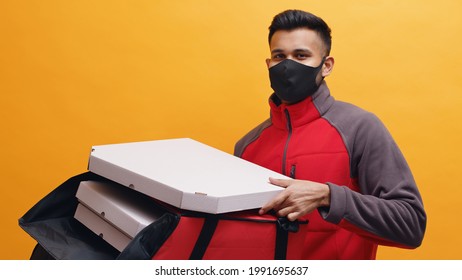 Delivery boy wearing black mask and carrying pizza boxes in his red parcel box. Concept of home delivery services during Covid-19. Isolated man with yellow background studio. . 