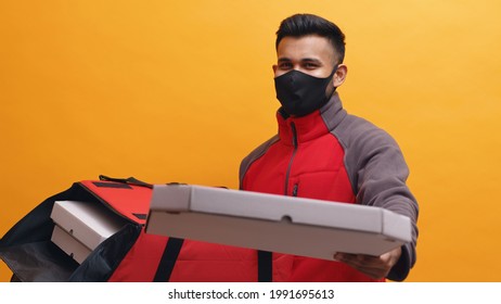 Delivery boy wearing mask carrying pizza boxes in his red parcel box. Holding a pizza box in his hand. Concept of home delivery services during Covid-19. Isolated man with yellow background studio.