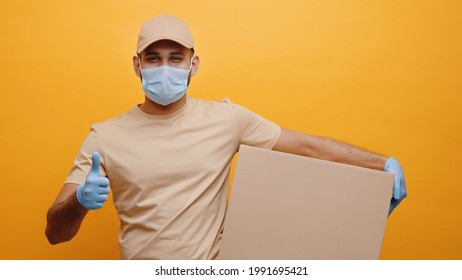 Delivery boy wearing mask and gloves holding a parcel box and making thumbs up sign. Concept of home delivery services during Covid-19. Isolated man with yellow background taking precautions. 