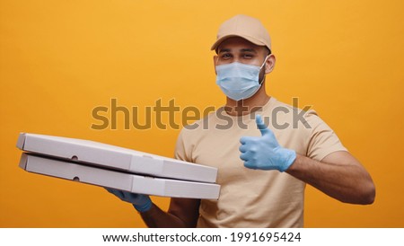 Delivery boy wearing mask and gloves holding pizza boxes making thumbs up sign and smiling. Concept of home delivery services during Covid-19. Isolated man with yellow background studio. 