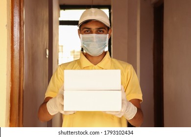 Delivery Boy Wearing Medical Face Mask And Gloves Holding Box Parcel At The Doorstep And Smiling.