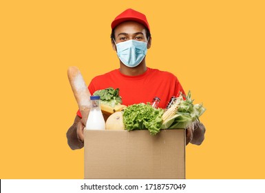 Delivery Concept - Handsome African American Delivery Man With Mask Carrying Package Box Of Grocery Food And Drink From Store. Isolated On Yellow Studio Background. Copy Space.