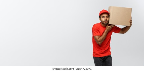 Delivery Concept - Portrait Of Curious African American Delivery Man Listen Inside A Box Package. Isolated On Grey Studio Background. Copy Space.