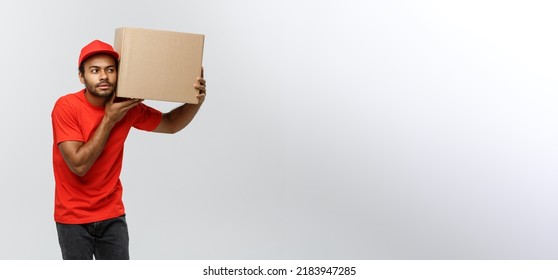 Delivery Concept - Portrait Of Curious African American Delivery Man Listen Inside A Box Package. Isolated On Grey Studio Background. Copy Space.