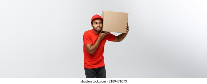 Delivery Concept - Portrait Of Curious African American Delivery Man Listen Inside A Box Package. Isolated On Grey Studio Background. Copy Space.