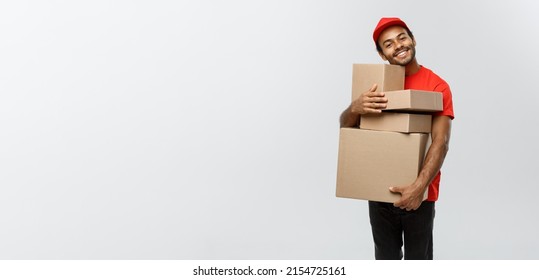 Delivery Concept - Portrait Of Happy African American Delivery Man In Red Cloth Holding A Box Package. Isolated On Grey Studio Background. Copy Space.