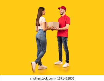 Delivery Courier Guy Giving Parcel Box To Woman Standing Over Yellow Studio Background. Post Packages Delivering, Parcels Transportation Service Concept. Full Length