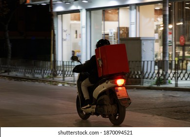 Delivery Courier On Unbranded Motorbike Speeding On City Road At Night. Illuminated View Of Male With Helmet Riding Motorcycle To Deliver Take Away Food Contained On A Red Box In Thessaloniki, Greece.