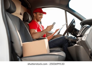 Delivery driver man wearing uniform, sitting inside van during the day, using smartphone while holding clipboard - Powered by Shutterstock