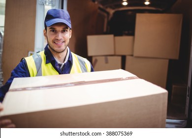 Delivery Driver Smiling At Camera By His Van Holding Parcel Outside The Warehouse