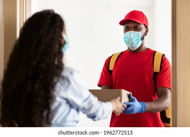 Delivery During Coronavirus Pandemic. Black Courier Guy Wearing Medical Face Mask And Protective Gloves Delivering Cardboard Box Order To Young Woman At Home, Standing At Door, New Normal